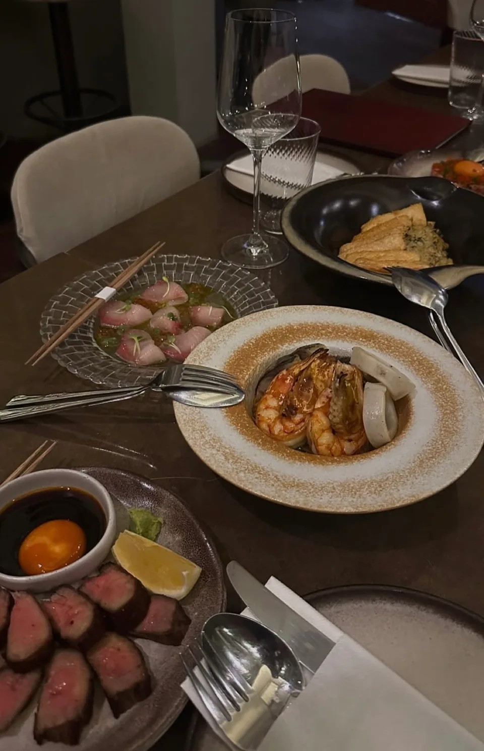 A shared dining table with seafood, sliced beef and wine glasses